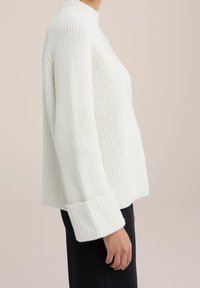 Bílý žebrovaný pletený svetr s vysokým límcem a širokými manžetami. Má volný střih, strukturovanou látku a zkrácený střih.