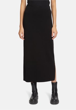 Frau trägt einen schwarzen, hoch taillierten Midirock mit einem seitlichen Schlitz und schwarzen, klobigen Stiefeletten, steht vor einem weißen Hintergrund.