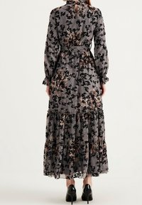 Maxi mekko läpinäkyvässä harmaassa kankaassa, jossa on musta kukkakuosi. Ominaisuuksiin kuuluvat pitkät hihat, kerroksellinen helma ja vyötäröllä solmittava vyö.