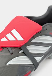 Scarpino da calcio grigio con una cintura superiore rosso brillante e bianca; presenta un materiale testurizzato, una forma elegante e accenti di design sorprendenti.