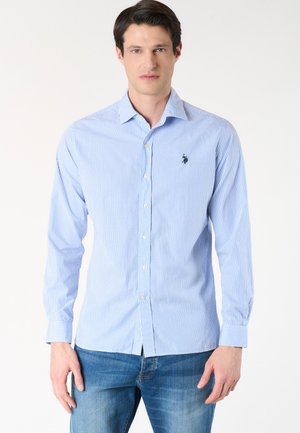 Uomo che indossa una camicia a quadri azzurri chiara con bottoni e maniche lunghe e jeans blu, in piedi su uno sfondo bianco.