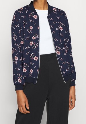 Femme portant une veste bomber bleu marine à motifs floraux sur un haut blanc et un pantalon noir, la veste partiellement dézippée.