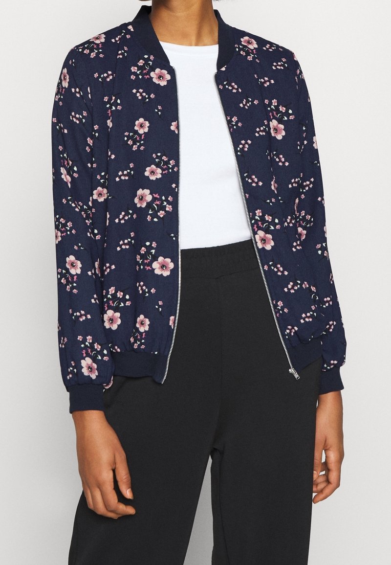 Femme portant une veste bomber bleu marine à motifs floraux sur un haut blanc et un pantalon noir, la veste partiellement dézippée.