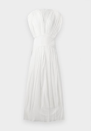 Žena stoji s rukom na kukovima, nosi dugu, lepršavu bijelu haljinu s dubokim V-izrezom i plisiranim materijalom na jednobijeloj pozadini.