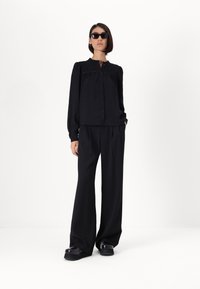 Black long-sleeve blouse with a button front, relaxed fit, and gathered detailing. Paired with wide-leg black trousers and chunky black shoes.