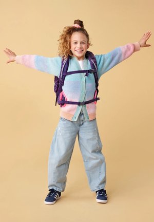 Young girl in pastel cardigan and loose jeans with a backpack, smiling and standing with arms outstretched on beige background.
