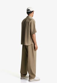 Relaxed-fit korte mouwen shirt en wijde broek in tan stof, gecombineerd met witte sneakers, getoond vanuit een achteraanzicht.