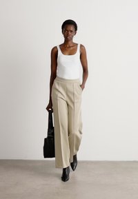 Witte tanktop, beige geplooide broek, zwarte haklaarzen en een zwarte tas. Het model staat voor een neutrale achtergrond.