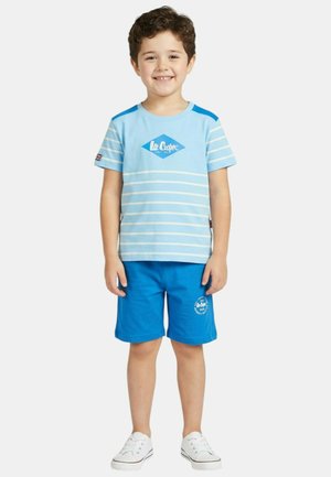 Jeune garçon souriant debout, portant un t-shirt rayé bleu clair Lee Cooper, un short bleu avec logo, et des baskets blanches sur fond blanc.