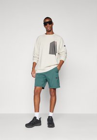 Bluza w jasnym kremowym kolorze z teksturowaną szaro-skalną kieszenią; zielone szorty; czarne sneakersy; duże czarne okulary przeciwsłoneczne. Prosty, swobodny styl.