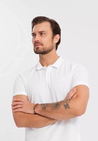 Valkoinen poolopaita, jossa on teksturoitu kangas, lyhyet hihat ja kaulus. Rinnassa on hienovarainen logo. Ihonväri näkyy käsissä, joissa on tatuointeja.