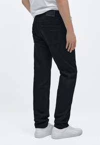 Černé džínové kalhoty z denimu s klasickým střihem, designem s pěti kapsami a jemnými šicími detaily, kombinované s bílými teniskami, pohled zezadu.