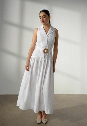 Vrouw draagt een mouwloze witte jurk met knopen, een brede rok en een decoratieve riem, staand op een vlakke vloer tegen een witte muur.