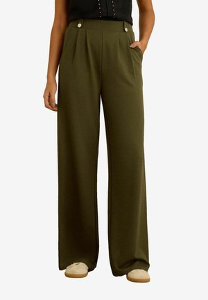 Naine kannab laia jalaga oliivrohelisi pükse, millel on voldid ja nööbid, kombineerituna beežide kingade ja musta varrukateta toppiga.