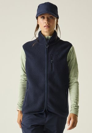 Woman wearing a navy fleece vest over a light green long-sleeve shirt, navy pants, and a navy cap, standing against a plain background.