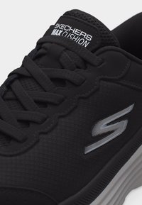 Μαύρο αθλητικό παπούτσι Skechers Max Cushion με μαύρα κορδόνια, υφή επάνω μέρος, ασημένιο λογότυπο στο πλάι και παχιά λευκή σόλα.