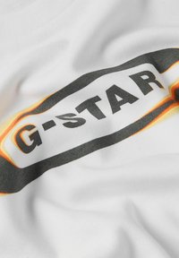 Bílá bavlněná tričko s černým a oranžovým logem G-STAR, s hladkým povrchem a volným střihem. Design zahrnuje zakřivené detaily.