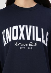 Tamsiai mėlynas džemperis su baltais užrašu "KNOXVILLE Leisure Club EST. 1982." Suapvalinta iškirptė ir minkšta medžiaga. Laisvas kirpimas.
