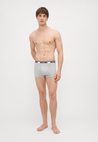 Boxers de algodão cinza com uma cintura preta da Moschino, apresentando um ajuste justo e uma textura suave. Modelo de pé descalço, sem camisa.