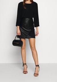 Haut noir à manches longues et jupe en cuir ajustée. Le mannequin tient un sac à main cylindrique noir. Des sandales à talons hauts noires à lanières complètent le look.