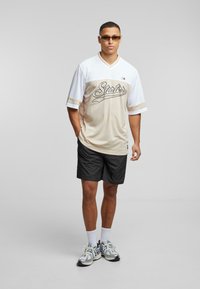 Beige and white short-sleeve jersey with "Starter" text, paired with black shorts. Model wears silver sunglasses and multicolor sneakers.
