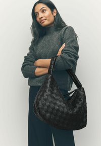 Černá tkaná kožená kabelka ve tvaru trojúhelníku, s robustním pleteným popruhem a zipovým uzávěrem, odpočívající vedle šedého outfitu.