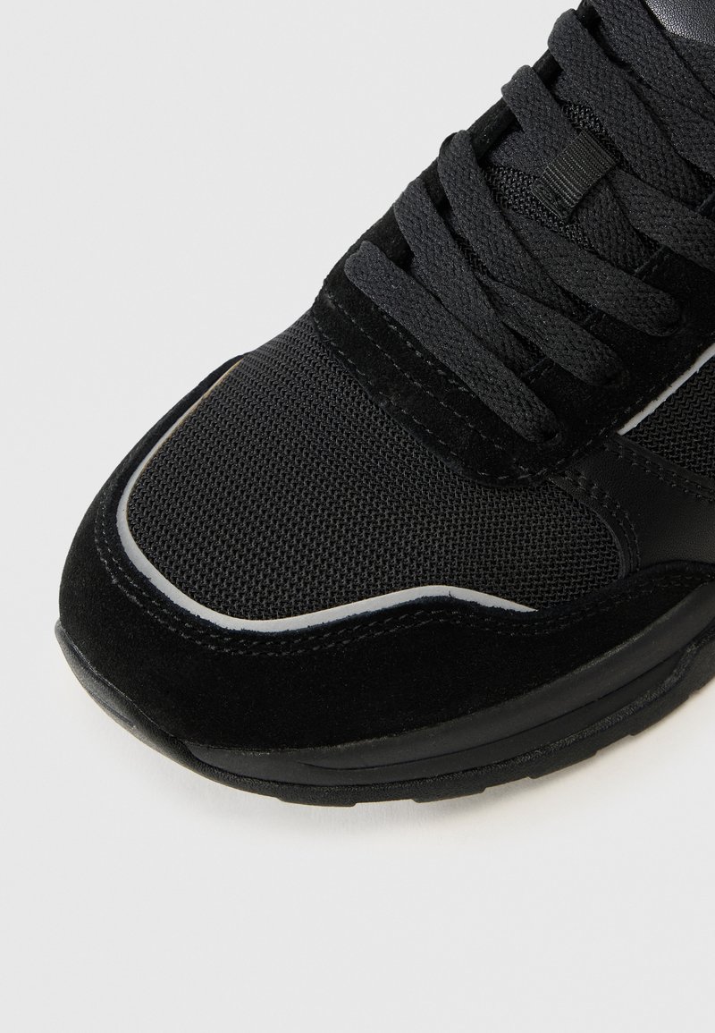 Schwarzer Sportschuh mit einem Obermaterial aus Mesh, Suede-Akzenten, flachen Schnürsenkeln und einer strukturierten Sohle, die mit weißen Akzenten und Nähten hervorgehoben wird.