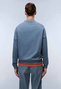 Lichtblauwe sweatshirt met ribgebreide manchetten en onderrand, met een oranje accent bij de hals, gedragen over grijze sportbroeken. Zachte textuur.
