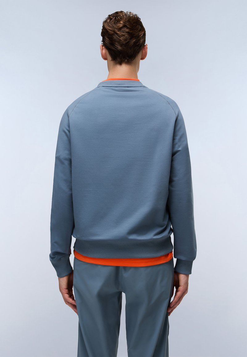 Hellblaues Sweatshirt mit gerippten Bündchen und Saum, mit orangefarbenem Akzent am Hals, getragen über grauen Sporthosen. Glatte Textur.