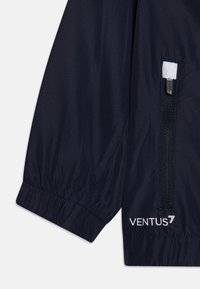 Veste coupe-vent marine avec poignets élastiques, poche à moitié zippée, détails en mesh et logo "VENTUS7" en blanc sur le bas.