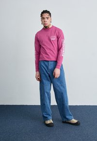 Camisa de algodón rosa de manga larga con el logo "OBEY"; pantalones vaqueros sueltos de mezclilla azul con diseño de patchwork; zapatos beige y negros.