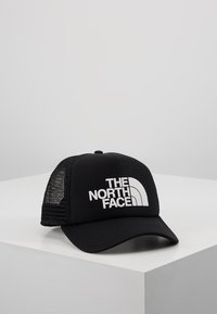 Casquette trucker noire en mesh avec une visière incurvée comportant le logo blanc "THE NORTH FACE" sur le devant, présentée sur une surface angulaire blanche.