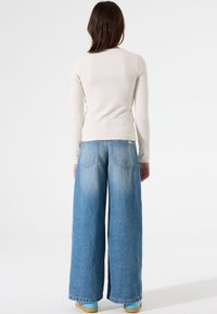 Beige top met lange mouwen en een aansluitend ontwerp, gecombineerd met lichtblauwe jeans met wijde pijpen. Opvallende rugpas en opgestikte zakken op de jeans.