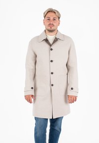 Cappotto di lana beige con colletto a punta, bottoni frontali e due grandi tasche. Presenta un accento marrone sul colletto e sui polsini con bottoni.