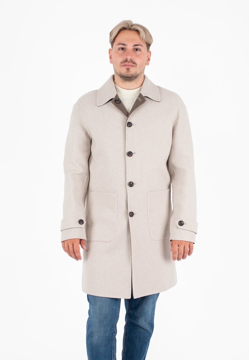 Cappotto di lana beige con colletto a punta, bottoni frontali e due grandi tasche. Presenta un accento marrone sul colletto e sui polsini con bottoni.