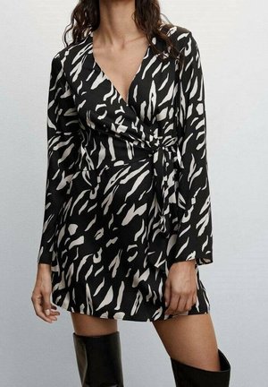 Femme portant une robe portefeuille à manches longues à imprimé zèbre noir et blanc, avec des bottes noires montantes jusqu'au genou, posant devant un fond uni.
