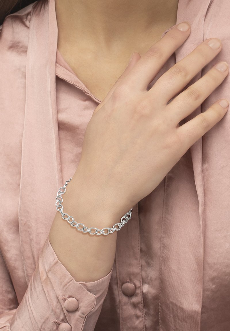 Bracciale a catena argento con collegamenti a forma di cuore intrecciati, indossato su un polso, con una blusa morbida rosa e texturizzata sullo sfondo.