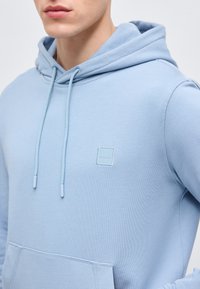 Κοντινό πλάνο ατόμου που φοράει ανοιχτό μπλε φούτερ με κορδόνια και μικρό τετράγωνο λογότυπο "BOSS" στο στήθος, χέρια στη μέση.