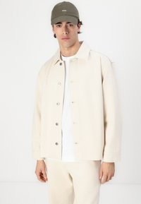Lys beige knappjakke i en strukturert stoff, brukt over en hvit skjorte, kombinert med en grønn caps med hvit brodering.