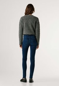 Suéter gris de punto con mangas texturizadas, combinado con jeans ajustados azul oscuro y zapatos negros, visto desde atrás.
