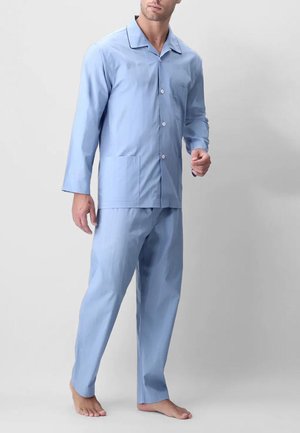 Uomo che indossa una camicia da pigiama azzurra a maniche lunghe con bottoni e colletto, abbinata a pantaloni larghi, in piedi a piedi nudi su sfondo neutro.