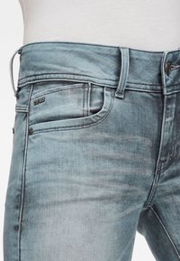 Světle modré džíny z denimu s úzkým střihem, vybledlou texturou, přední kapsou a kovovým knoflíkem s logem 'RAW' poblíž pasu.