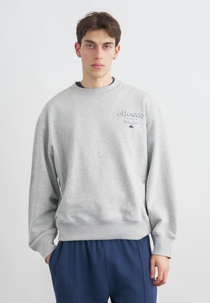 Jeune homme aux cheveux courts et bruns portant un sweat-shirt gris clair de la marque ellesse et un pantalon bleu marine, debout devant un fond blanc uni.