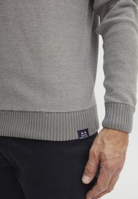 Maglione orizzontale a strisce grigie con polsini e orlo a costine. Presenta un'etichetta con logo nella parte inferiore con accenti rossi, bianchi e blu.