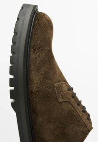 Chaussure en daim marron avec un bout arrondi, surface texturée et semelle épaisse noire présentant un motif de semelle et des détails de lacets sur le côté.