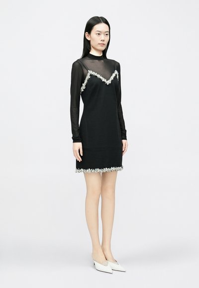 Melna kleita ar caurspīdīgu melnu garām piedurknēm, ko rotā ziedu dekorācijas pie izgriezuma un apakšmalas. Modelis valkā baltus kurpes uz papēžiem.