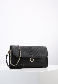 Černá kabelka na psaní s texturovaným klopněním, zlatým kroužkem a odnímatelným zlatým řetízkovým popruhem, ležící na bílé ploše na světlem pozadí.