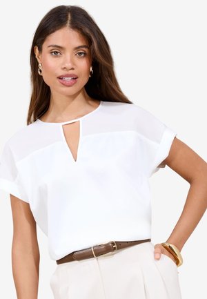 Nő hosszú barna hajjal, fehér rövid ujjú blúzzal, elöl kivágott részlettel, bézs nadrággal, barna övvel, arany karika fülbevalókkal és arany karkötővel.