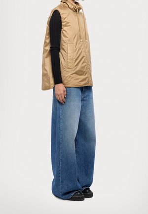 Person trägt eine hellbraune ärmellose Kapuzenweste, ein schwarzes geripptes Langarmoberteil, weit geschnittene blaue Jeans und schwarze Schuhe, steht vor einem weißen Hintergrund.