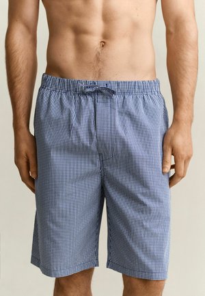 Homme portant un short à cordon de serrage à carreaux bleu et blanc, debout avec les bras détendus le long du corps devant un fond uni.